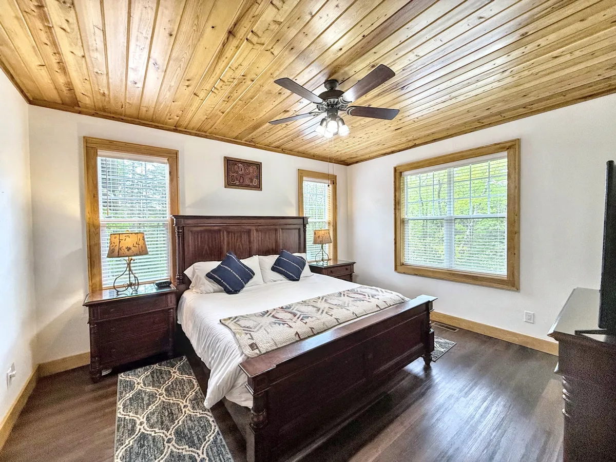 A serene bedroom featuring a plush bed with decorative pillows, a wooden ceiling, and large windows that welcome abundant natural light. Nightstands with lamps flank each side of the bed, creating a tranquil ambiance ideal for rest and relaxation.