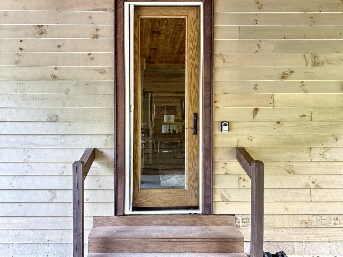 This image features the home's exterior front door, wooden steps, and rustic siding, offering a glimpse of the welcoming entrance surrounded by natural elements. The shot captures the connection between the indoors and the lush outdoor setting.