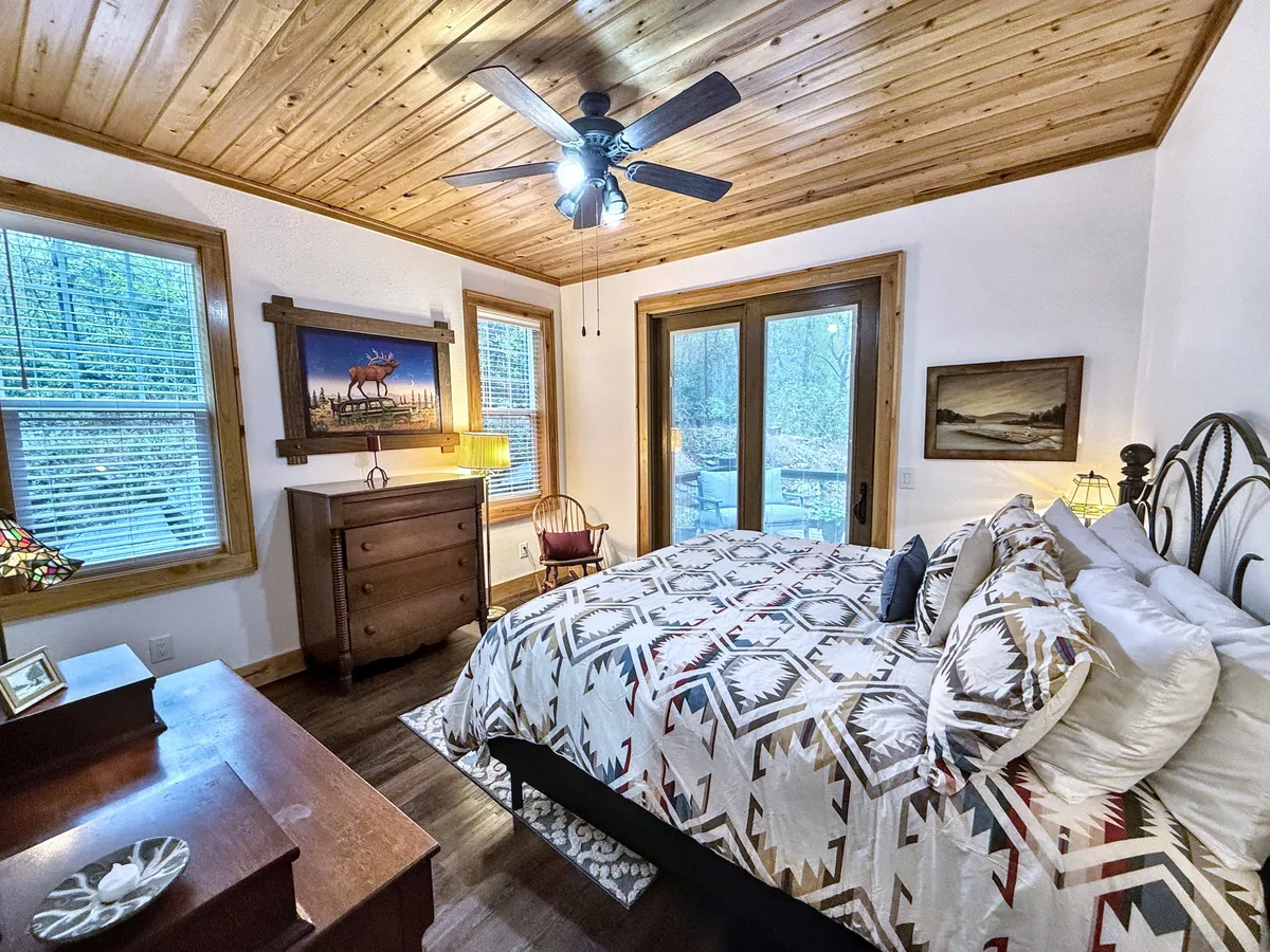 A tranquil bedroom featuring patterned bedding, a wooden dresser, and large windows that frame picturesque outdoor views. Subtle artwork and cozy touches create a serene retreat, while natural light pours in, providing an inspiring backdrop for reading or morning routines.