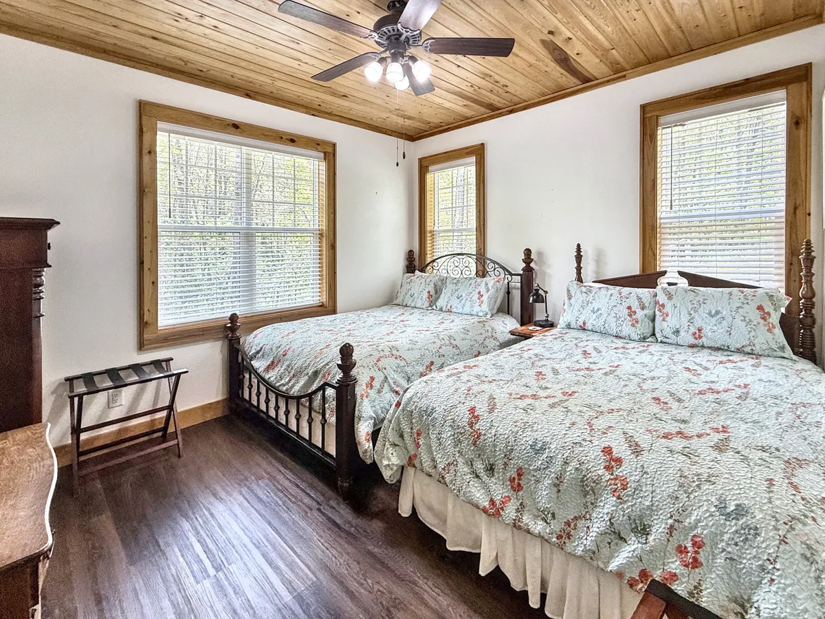 A bright, inviting room with two beds adorned in floral quilts, large windows flooding the space with natural light, and a vintage dresser. The windows offer glimpses of the outdoors, enhancing the sense of connection to the surrounding landscape.