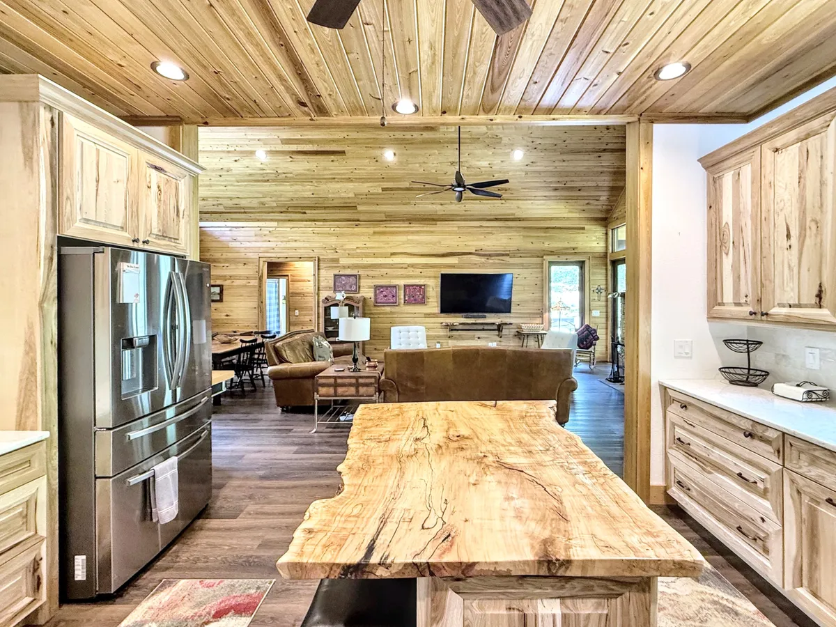 A spacious, open-concept kitchen and living area featuring a central island with bar seating, a modern refrigerator, and comfortable lounge spaces under a warm wooden ceiling. Large windows suggest scenic views and plenty of natural light, while the layout invites both productivity and relaxation.
