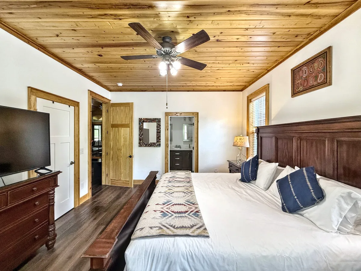 A cozy king bedroom with a comfortable bed, wooden ceiling, television, mirror, and decorative art. The room is filled with natural light, making it perfect for waking up refreshed or relaxing with a favorite show.