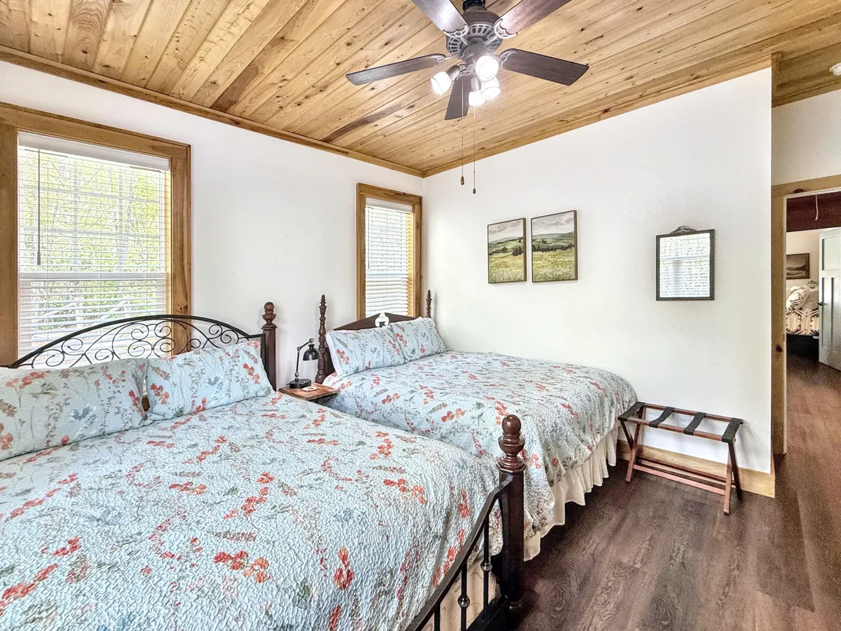 A spacious guest room featuring two queen-sized beds adorned with floral quilts, a wooden ceiling fan, and framed artwork on the wall. The space feels bright and inviting, ideal for sharing with family or friends.
