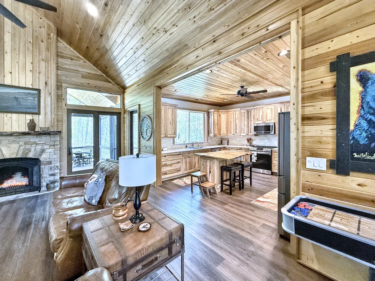 The image features a cozy living area with a prominent stone fireplace, wooden cabinetry, large windows revealing glimpses of the outdoors, a kitchen island, and comfortable seating. The warm tones and inviting atmosphere evoke the feeling of gathering indoors during cooler months or enjoying the view of seasonal changes from a cozy vantage point.