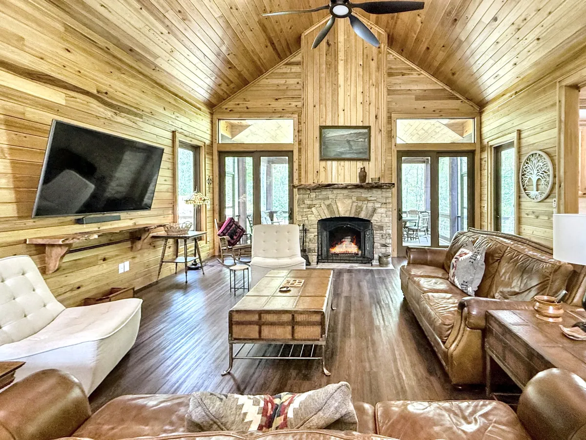 This image features a cozy living space with a wood-burning fireplace as the focal point, surrounded by sofa seating and a coffee table. The wood-paneled walls and large windows allow natural light to fill the room, creating a warm and inviting atmosphere ideal for gatherings or quiet relaxation.