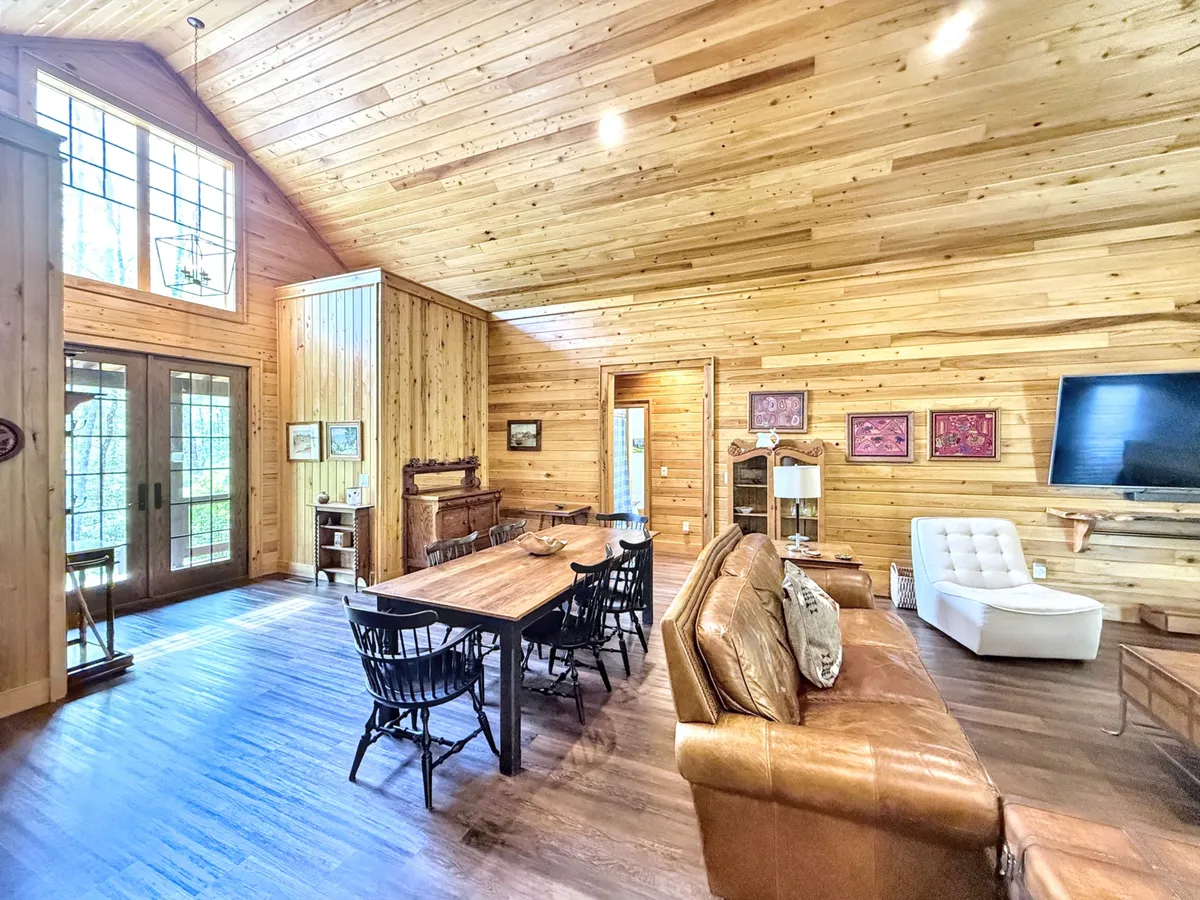 An open-plan space combining a large wooden dining table, a comfortable leather sofa, and large windows that flood the area with sunlight. The setup encourages effortless flow between living, dining, and relaxation zones.