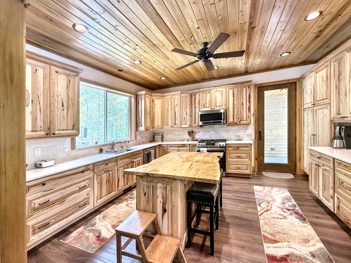 An open kitchen scene with a large wooden island, natural wood cabinetry, a large window letting in abundant light, stools, and a patterned rug. The setting feels welcoming, perfect for cooking together, sharing meals, or enjoying late-night snacks after seasonal adventures.
