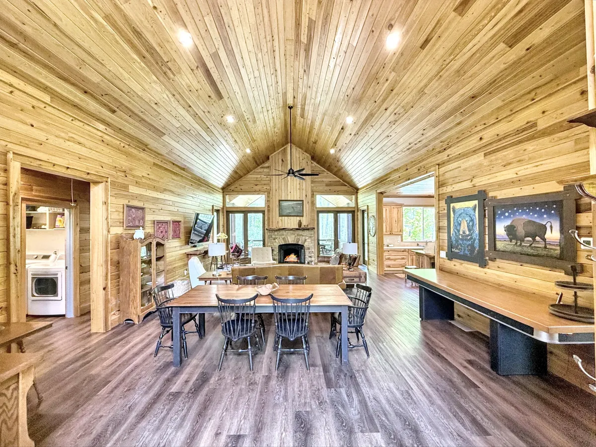 A large dining table set in a cozy, wood-accented room with a stone fireplace, wooden ceiling, and expansive windows showcasing the surrounding forest. The artwork and warm lighting create an inviting atmosphere ideal for family gatherings or romantic dinners. The visual connection to the outdoors through the windows enhances the woodland dining theme.