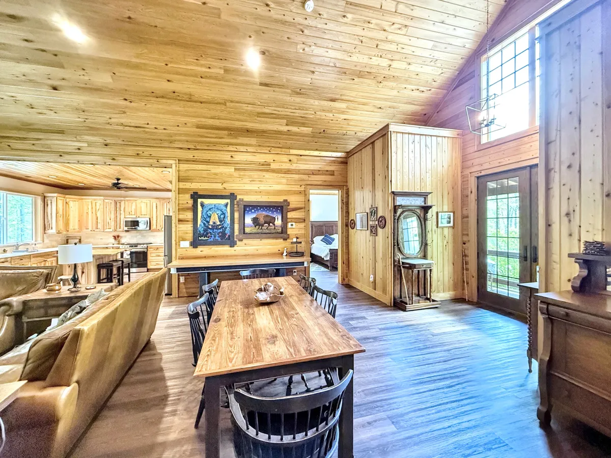 A warm, inviting dining and living area with a large dining table, comfortable sofa, striking artwork, and expansive windows. The space is bright and airy, ideal for family gatherings, holiday meals, or simply relaxing while enjoying the natural beauty outside.