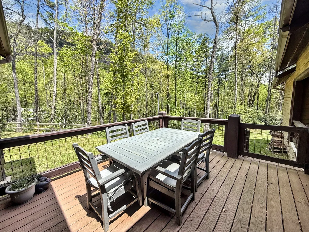 A wooden dining table sits on a spacious deck surrounded by lush green trees, with sturdy deck railings that create a sense of enclosure while still allowing for immersive views of the landscape. Comfortable seating invites guests to linger and enjoy the serene ambiance, with hints of nature peeking through at every angle.