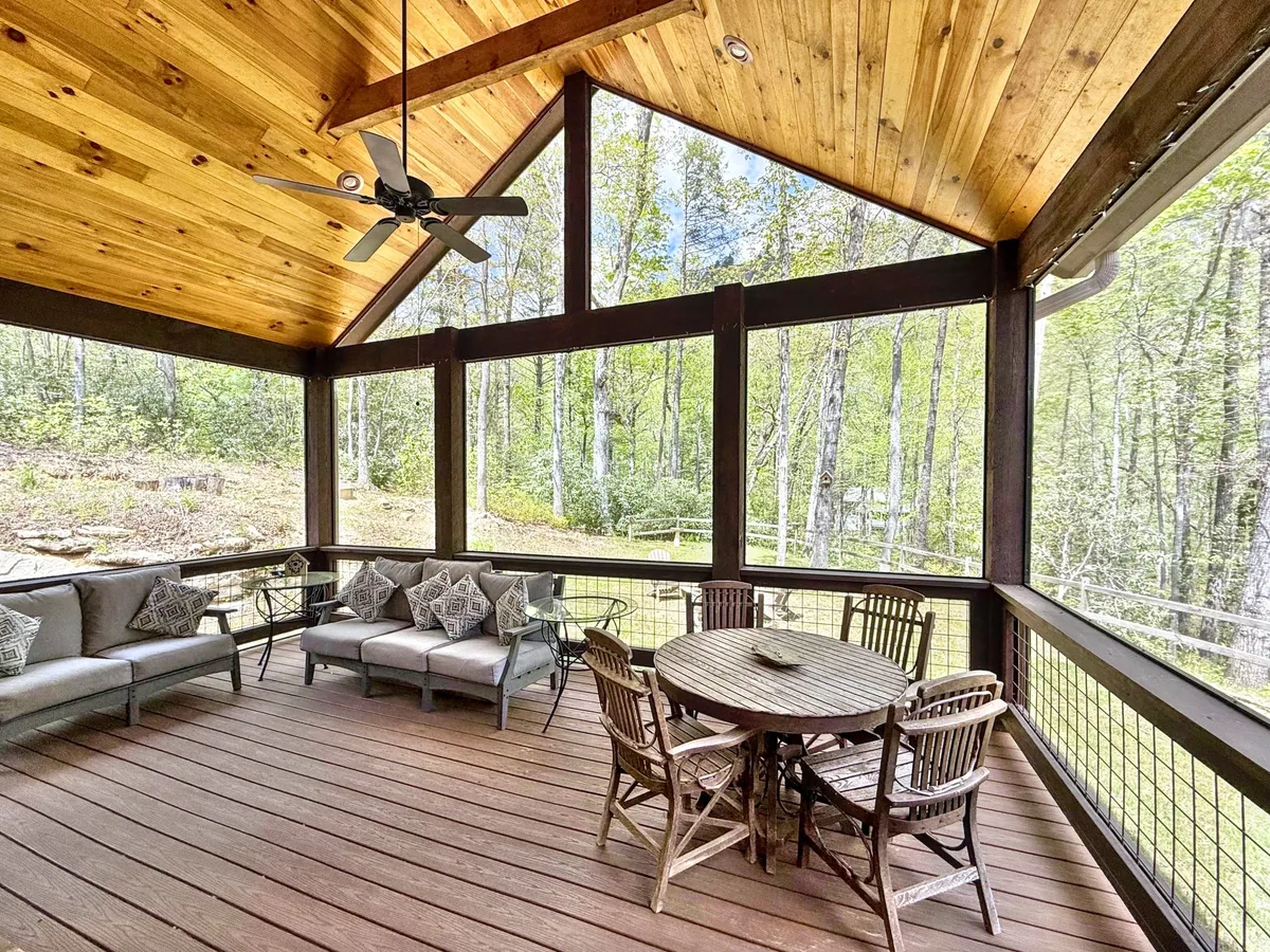 The image showcases the exterior of a charming single-family cabin surrounded by lush greenery, featuring a spacious outdoor seating area with a round dining table under a warm wooden ceiling. The setting is tranquil, perfect for gatherings, dining al fresco, or relaxing while enjoying the serene forest views and dappled sunlight filtering through the trees.