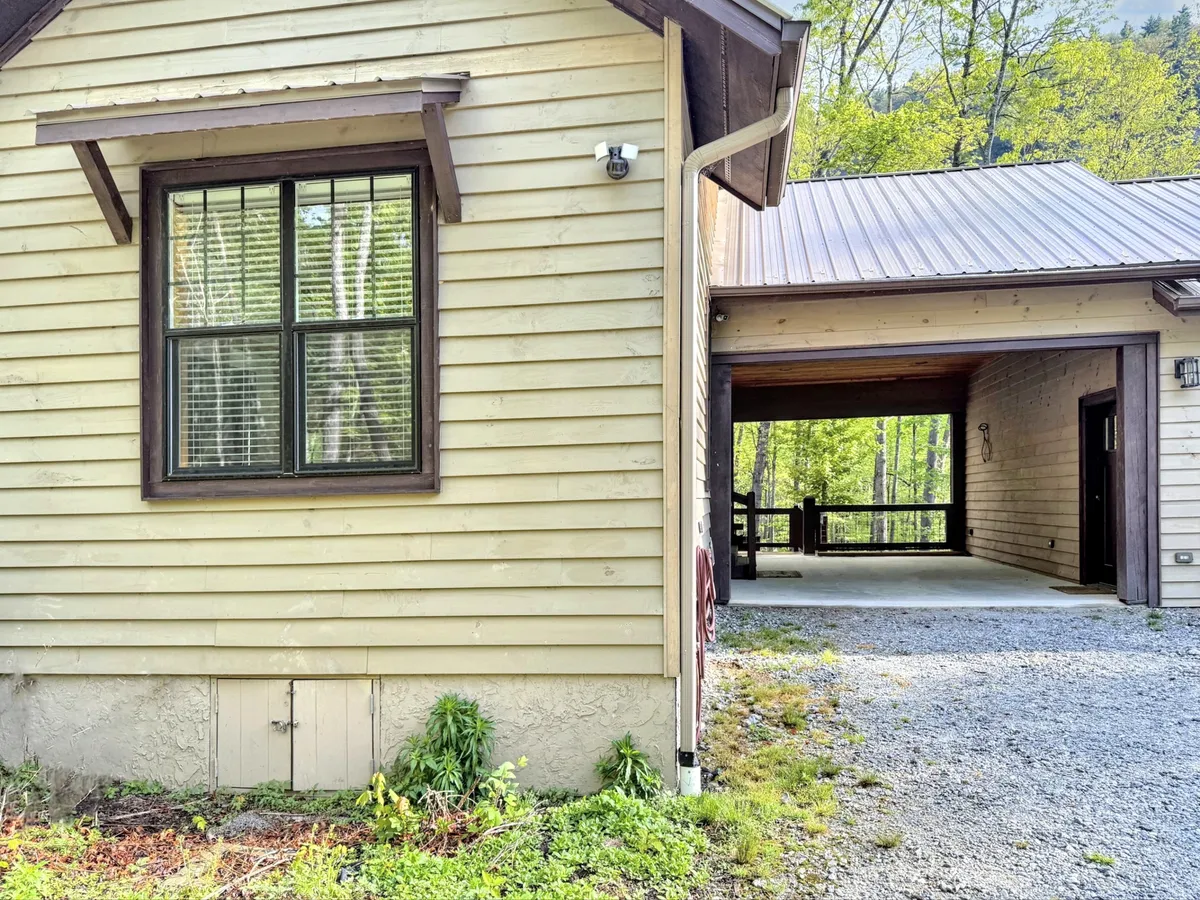 An exterior shot featuring a window with a grid design and the entrance to a carport, surrounded by natural foliage. The space feels inviting and tranquil, with hints of morning light creating a welcoming atmosphere, suggesting an easy start to a day of exploration.